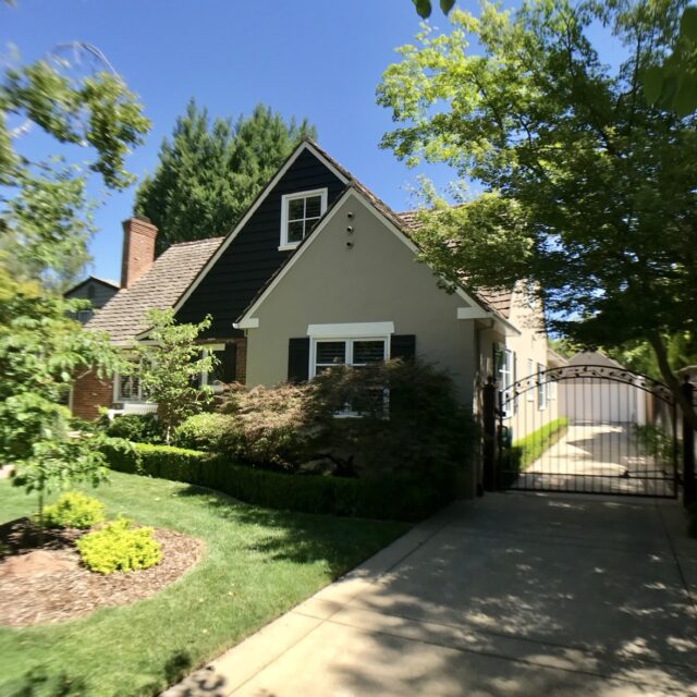 Exterior house painting in Sacramento, CA by Baldwin Custom Painting — looking up at a dark-shingled gable with a small white window, tan walls, and a brick chimney framed by tall trees